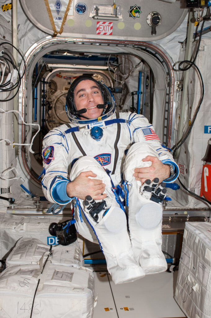 NASA Astronaut Chris Cassidy floating weightlessly in a tucked position inside an International Space Station module. Wearing a Russian Sokol pressure suit during the Expedition 63 mission, he illustrates the "fun of being there" and the physical adaptation required to lead in a zero-G environment.