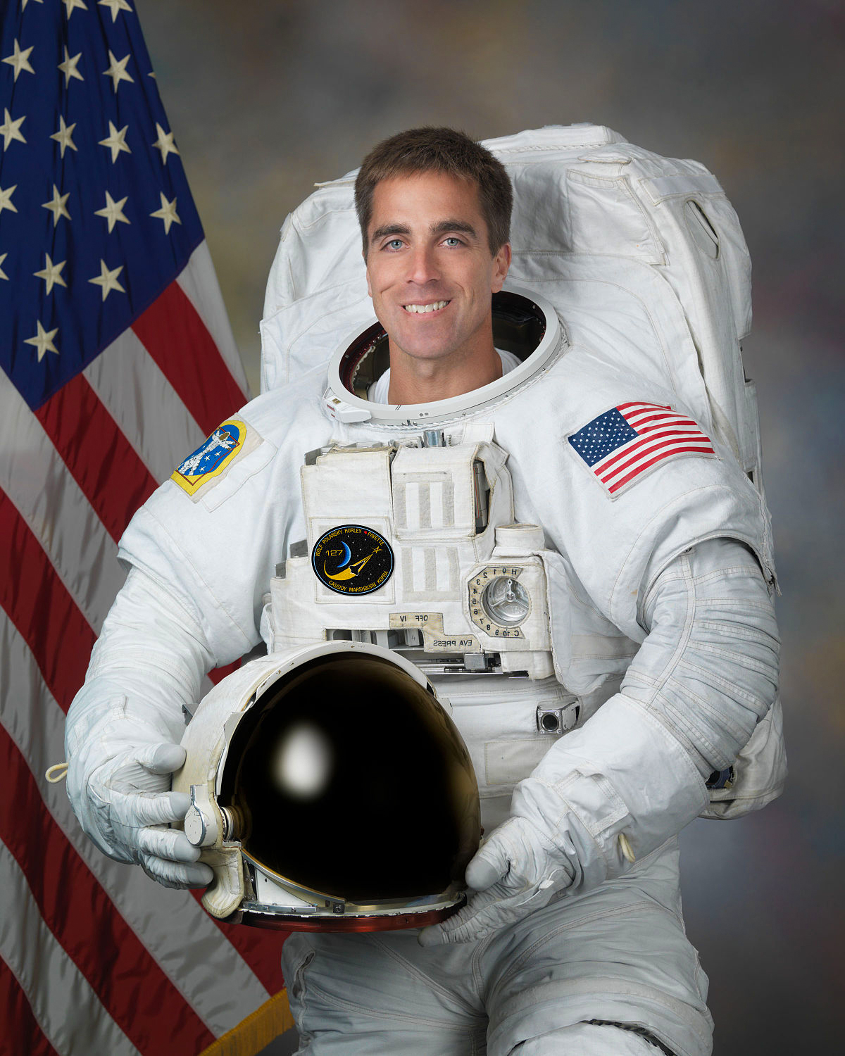 Official NASA studio portrait of Captain Chris Cassidy wearing his white Extravehicular Mobility Unit (EMU) spacewalking suit. He is smiling while holding his helmet, with the American flag prominently displayed in the background.